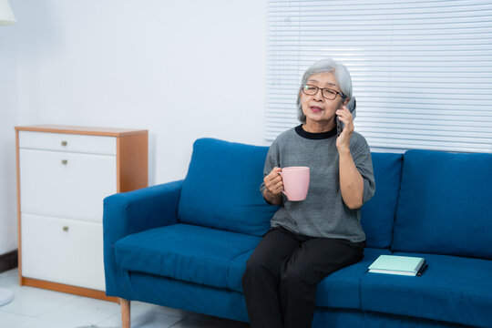 Elderly woman sits on a blue sofa, holding a pink mug and talking on the phone. A notebook rests beside her, suggesting a relaxed and casual conversation at home