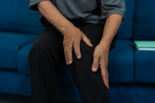 Close-up capturing a senior man experiencing knee pain while sitting on a sofa at home, with hands gripping his painful knee, highlighting the challenges of healthcare for the elderly