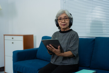Elderly asian woman wearing headphones is using a tablet while sitting on a sofa in her living room, enjoying technology and staying connected in retirement