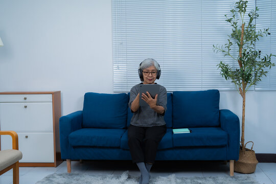 Relaxed senior asian woman using digital tablet and wearing headphones while sitting on blue sofa in living room at home, enjoying technology and online entertainment
