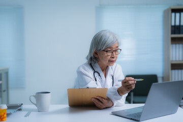 Experienced Asian female doctor wearing eyeglasses and lab coat is having video call with patient. Providing online consultation using laptop and taking notes on clipboard in her bright modern office
