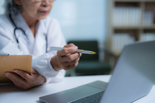 Senior female doctor holding a pen and clipboard, explaining a diagnosis during an online consultation while using her laptop in a well-equipped office setting