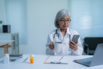 Experienced senior doctor in a white coat multitasking with a smartphone and coffee in a modern...