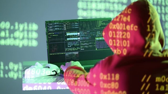 A masked individual in a hoodie types on a laptop while hexadecimal digits and system code illuminate their workspace. A white mask lies nearby, representing anonymity in the digital shadows