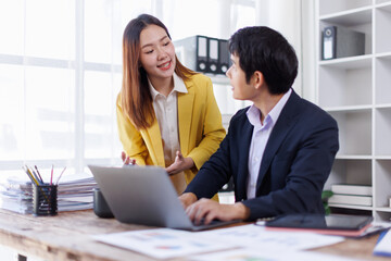Two happy asian employees working together using computer planning project. business woman consulting teaching young employee looking at laptop sitting at desk in office
