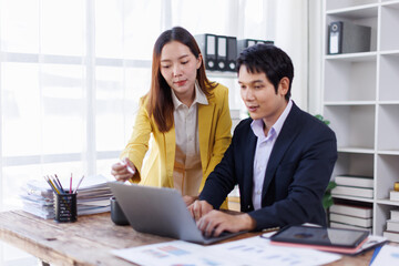 Two happy asian employees working together using computer planning project. business woman consulting teaching young employee looking at laptop sitting at desk in office
