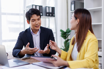 Two happy asian employees working together using computer planning project. business woman consulting teaching young employee looking at laptop sitting at desk in office
