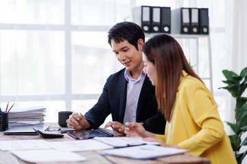Two happy asian employees working together using computer planning project. business woman consulting teaching young employee looking at laptop sitting at desk in office
