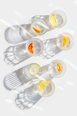 Crystal glasses with citrus slices in water on white background with rainbow reflections at sunlight, modern, minimal composition with glass cups filled with water and citrus fruit, as lemon orange