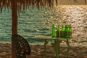 Green beer bottles are sitting on a table on a beach at sunset, under a straw umbrella, creating a relaxing and refreshing scene