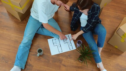 Cheerful man and woman sitting on the floor in new house with coffee and home project discussing interior design and reconstruction. Family moving in new home. Top view. Slow motion