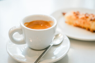 A white cup of espresso on a plate, accompanied by a blurred cream cake in the background, capturing a simple pleasure of life