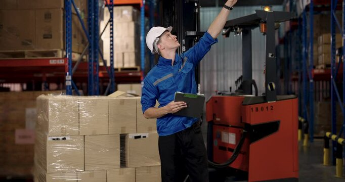 Caucasian male warehouse supervisor holds clipboard and points upward during forklift check while reviewing storage area logistics and documenting observations in cargo environment with industrial