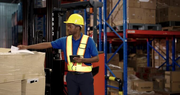 African warehouse worker wearing helmet and safety vest holds digital tablet while reaching toward document on stacked boxes during supply checking and cargo verification near forklift machine
