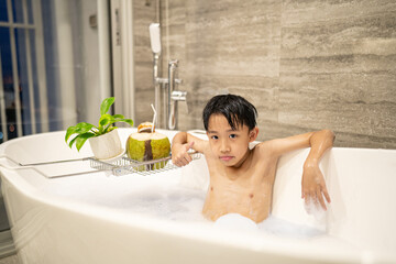 Asian boy relaxing in bathtub with coconut drink and foam bubbles in modern bathroom