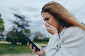 Worried woman looking at phone screen outdoors with anxious expression, emotional stress, casual...