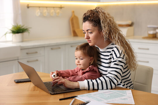 Single mother holding her daughter while working on laptop at wooden table in kitchen