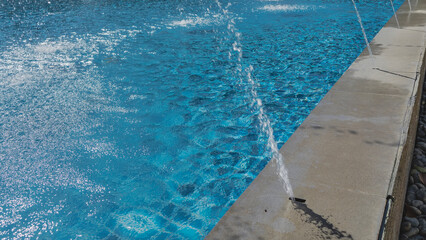 A fragment of an outdoor swimming pool. Blue tiled bottom, clear water. Highlights, sparks, ripples on the surface. There are fountain streams around the perimeter. Close-up