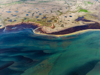 Aerial shot of a coastal plain filled with blue ponds, a winding river, and colorful sediments flowing into the turquoise sea, forming a natural abstract masterpiece.