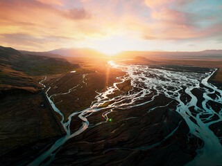 Golden sunset over a complex network of glacial rivers flowing across a dark Icelandic plain, with intense warm light and vibrant reflections on the water.