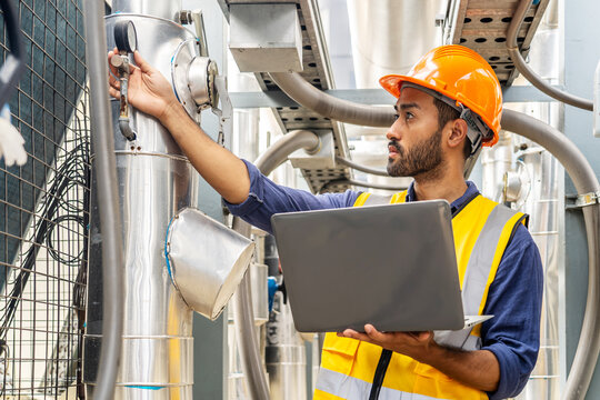 Male technical foreman in safety uniform inspects maintenance work work holding a laptop to look at plumbing and electrical systems on the roof of a building, Industrial background