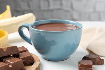 Tasty chocolate milk in cup, pieces and banana on white wooden table, closeup