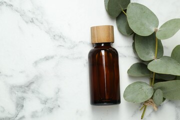 Bottle of essential oil and eucalyptus leaves on white marble table, flat lay. Space for text