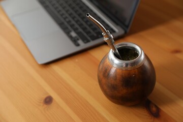 Traditional yerba mate tea and laptop on wooden table, closeup