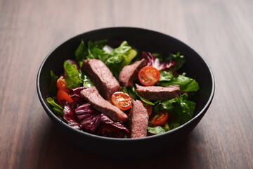 Steak salad with cherry tomatoes and mixed greens in black bowl on walnut wood background