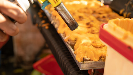 treet food chef using twin torches to dim sum caramelize melted cheese on a tray of baked food. Close-up of culinary flame technique, cheese topping, and gourmet snack preparation.