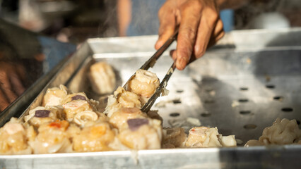 Close-up of a hand using tongs to pick steamed dim sum from a metal tray, with natural lighting and...