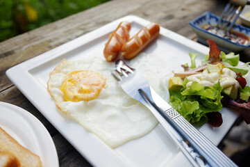 Close up hot dog and fried egg on white dish with fork and knife on wooden table for breakfast in the morning in garden of cafe