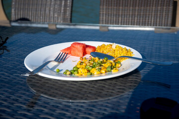 Morning breakfast includes pieces of red melons, poha, chickpeas, and corn served on a white plate. A fork and spoon are placed on the table for convenience.