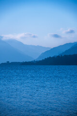 Blue mist covers the mountains, while a blue lake reflects the scenery. A cloudy sky adds depth to the view of the multi-layered mountains surrounding the area.