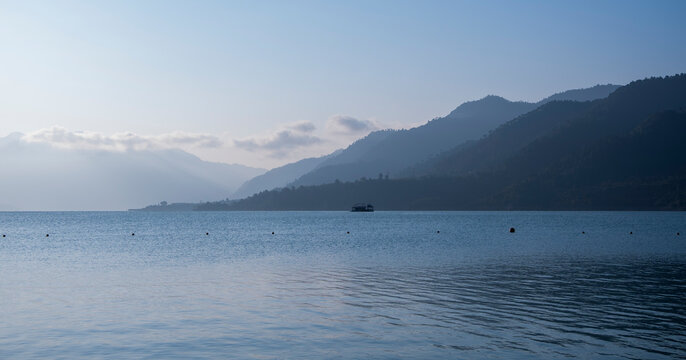 Blue mist shrouds the mountains in morning light, with a blue lake below. Light reflects on the waves, and a floating hotel is visible far away under a cloudy sky.