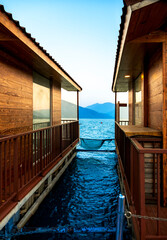 Blue lake water glistens below floating wooden huts, while blue mist covers the mountains on the other side. Yellow sunlight enhances the stunning landscape.