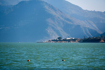 A green lake reflects the blue mist-covered mountains, while a small town rests on its shore. The...