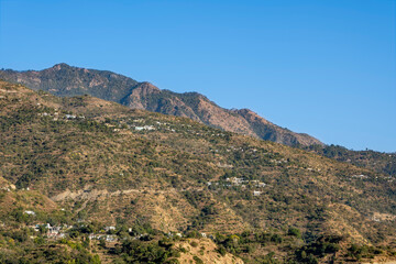 The sun shines brightly on the mountain, casting a yellow light on green-covered slopes with small towns nestled within. The picturesque view is inviting.