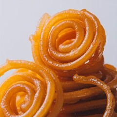 The image showcases a stack of traditional Indian sweets, jalebi, with their golden-orange color and spiral shapes, glistening with sugar syrup and set against a plain white background.