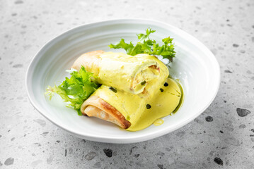 Two rolled crepes are drizzled with a rich yellow sauce and garnished with fresh greens, elegantly arranged in a white bowl. The dish showcases a delightful culinary presentation.
