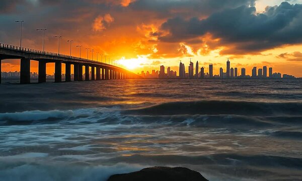 Sunset over city skyline with bridge and crashing waves in foreground