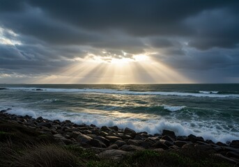 The sun breaks through dramatic clouds, casting light over a stormy ocean and rocky shore.