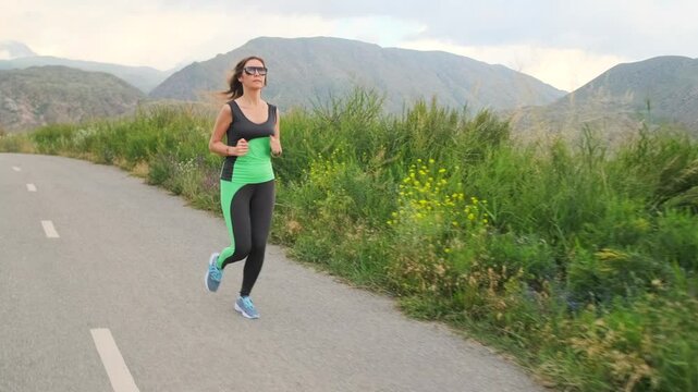 A young woman in sportswear runs at a steady pace along a paved road with a gentle decline. Her regular training helps reshape her skinny fat body type, set against rolling hills and green foothills.