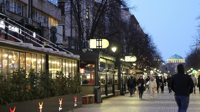 SOFIA, BULGARIA - FEBRUARY 11, 2025: Evening view of Vitosha Boulevard with blurred pedestrians, streetlights, cafes decorated with festive string lights and lighted St. Nedelya Church in background
