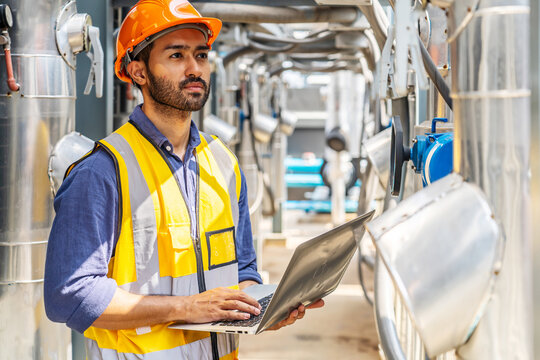 Male technical foreman in safety uniform inspects maintenance work holding a laptop to look at plumbing and electrical systems on the roof of a building, Industrial background