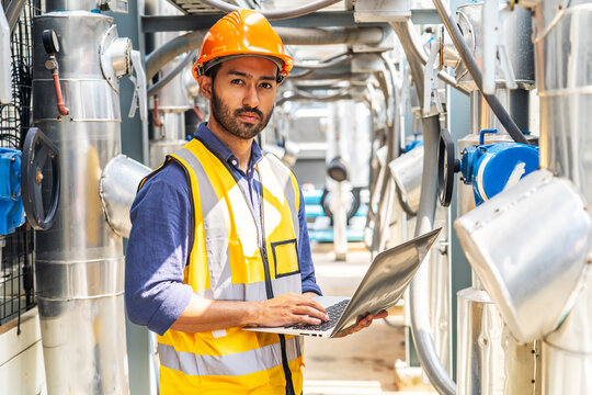Asian male technical foreman in safety uniform inspection maintenance work holding a laptop to look at plumbing and electrical systems on the roof of a building, look confidently at the camera