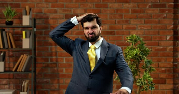 Showing thumbs down with disappointed facial expression, Indian Asian businessman or CEO in suit and beard expressing dissatisfaction or rejection in cozy modern office against terracotta brick wall