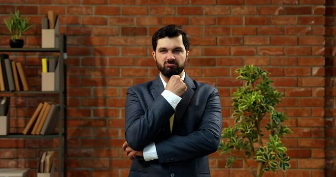 Showing thumbs down with disappointed facial expression, Indian Asian businessman or CEO in suit and beard expressing dissatisfaction or rejection in cozy modern office against terracotta brick wall