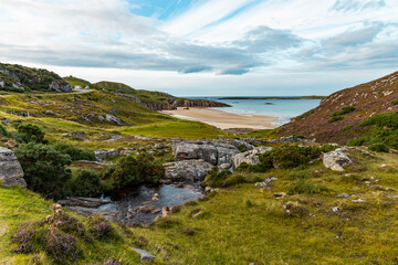 Ceannabeinne Beach shines under a clear summer sky. Crystal-clear waters, golden sand with a heart drawn on it, and lush green grass create a pristine and romantic coastal escape.

