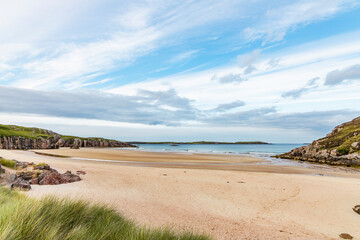 Ceannabeinne Beach shines under a clear summer sky. Crystal-clear waters, golden sand with a heart drawn on it, and lush green grass create a pristine and romantic coastal escape.

